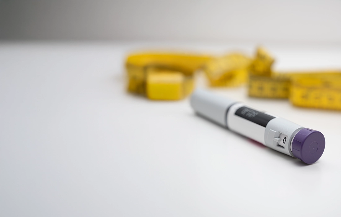 Zbliżenie pena insulinowego z fioletową nasadką leżącego na białej powierzchni, z żółtą taśmą mierniczą rozmytą w tle.