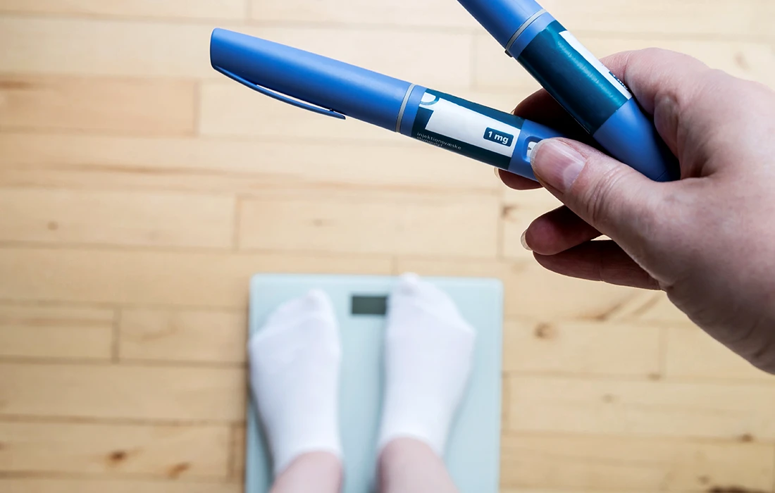 Ręka trzyma dwa niebieskie peny insulinowe nad wagą łazienkową, a na wadze widoczne są stopy osoby w skarpetkach.