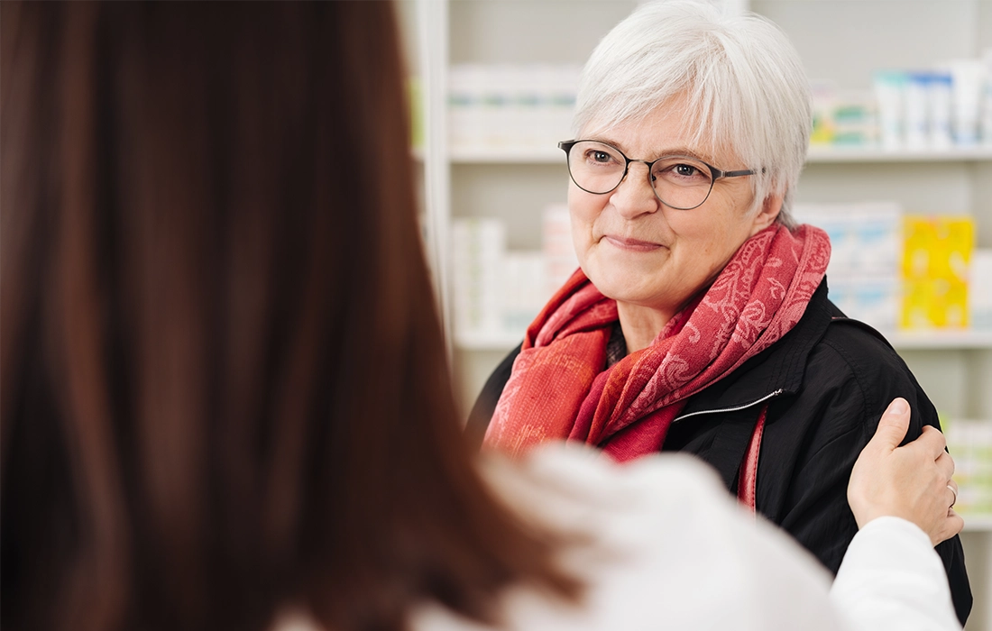 Starsza kobieta z krótkimi białymi włosami i okularami, ubrana w czerwony szalik, uśmiecha się, rozmawiając z inną osobą, która trzyma rękę na jej ramieniu.