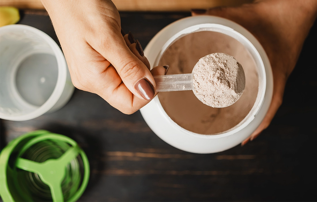 Ręka trzymająca miarkę czekoladowego białka w proszku nad otwartym pojemnikiem z zieloną pokrywką na ciemnej powierzchni.