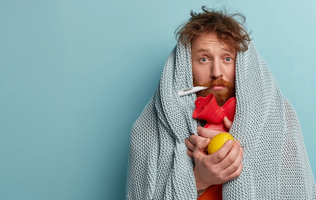 Chory mężczyzna owinięty w koc trzyma butelkę z gorącą wodą i cytrynę, z termometrem w ustach, stojąc na niebieskim tle.