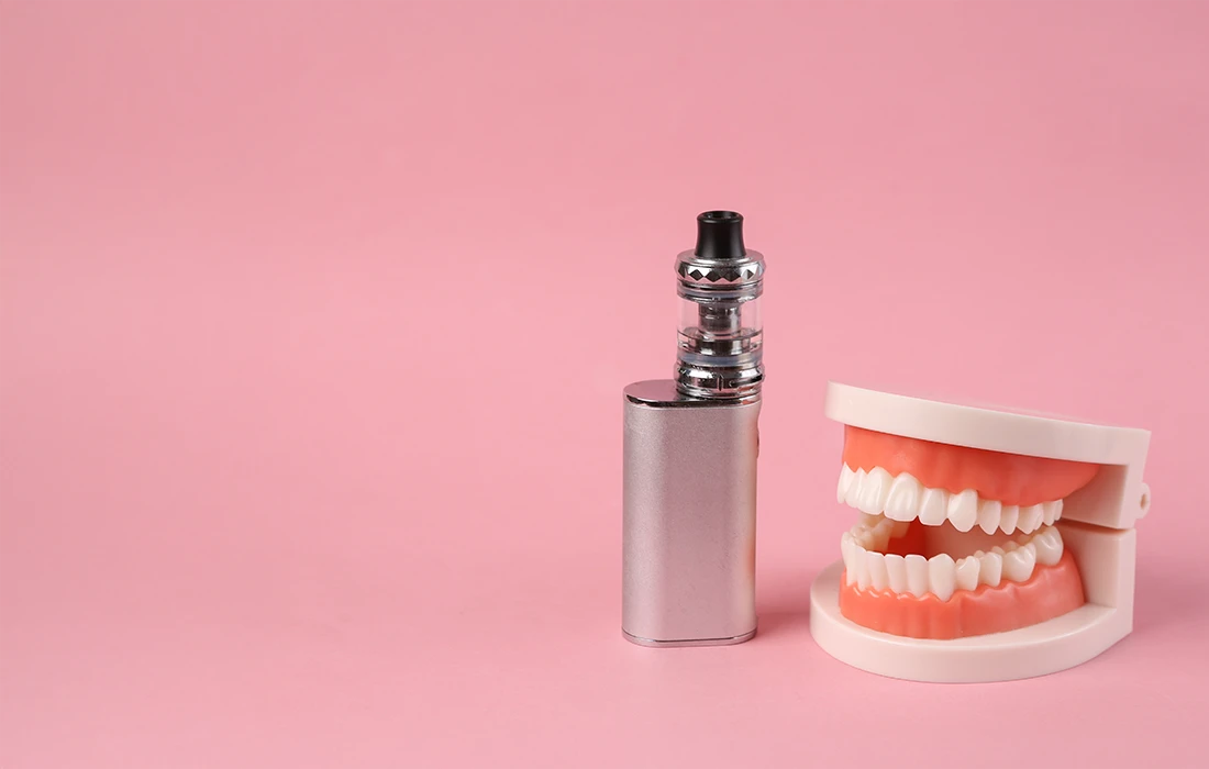 Urządzenie vape i model ludzkich zębów są umieszczone obok siebie na różowym tle.