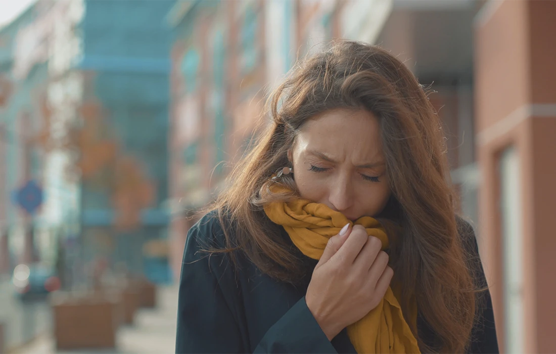 Kobieta na zewnątrz zakrywa usta żółtym szalikiem podczas kaszlu, w tle rozmazana ulica miasta.