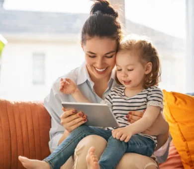 Uśmiechnięta kobieta i młoda dziewczyna siedzą na kanapie, patrząc razem na tablet. Kobieta trzyma dziewczynkę na kolanach, a oni wydają się szczęśliwi i zaangażowani.