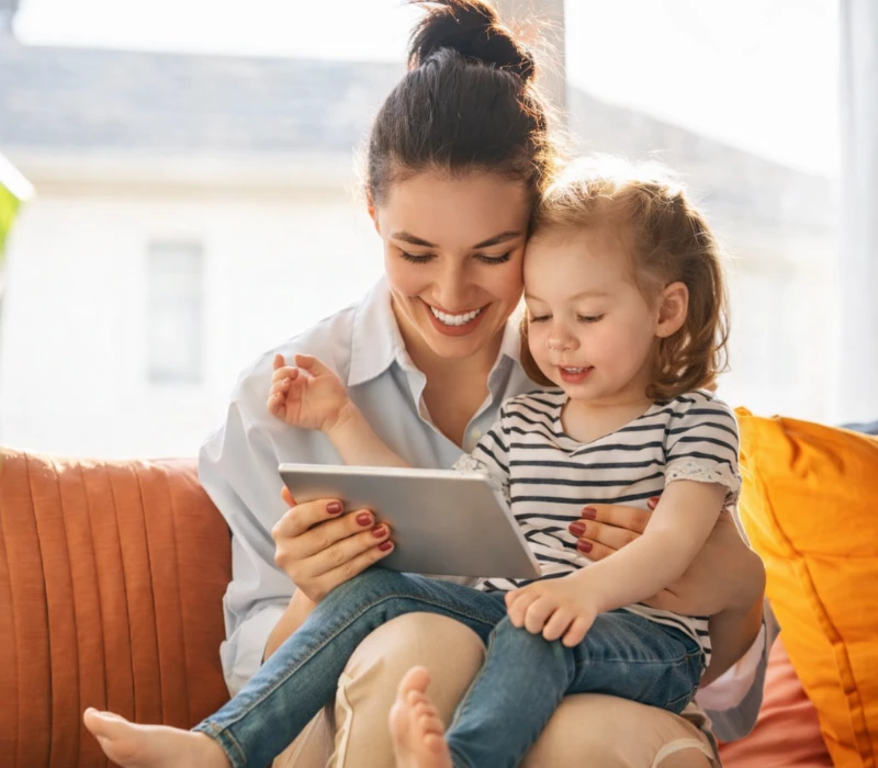 Uśmiechnięta kobieta i młoda dziewczyna siedzą na kanapie, patrząc razem na tablet. Kobieta trzyma dziewczynkę na kolanach, a oni wydają się szczęśliwi i zaangażowani.