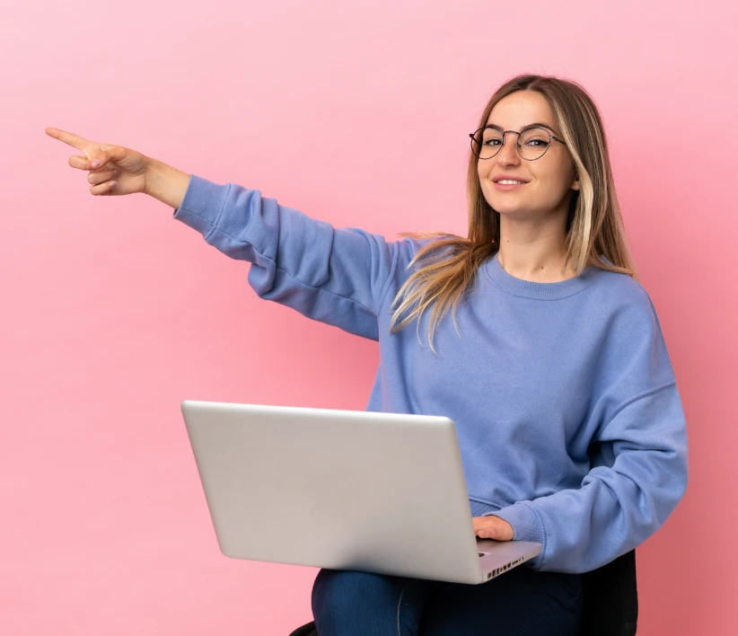 Kobieta w okularach i niebieskim swetrze siedzi z laptopem na kolanach, uśmiecha się i wskazuje w lewo na różowym tle.