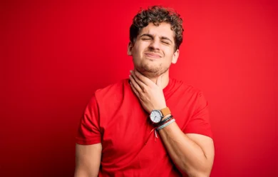 Mężczyzna w czerwonej koszuli stoi na czerwonym tle, dotykając swojego gardła z wyrazem bólu, sugerującym ból gardła lub dyskomfort.